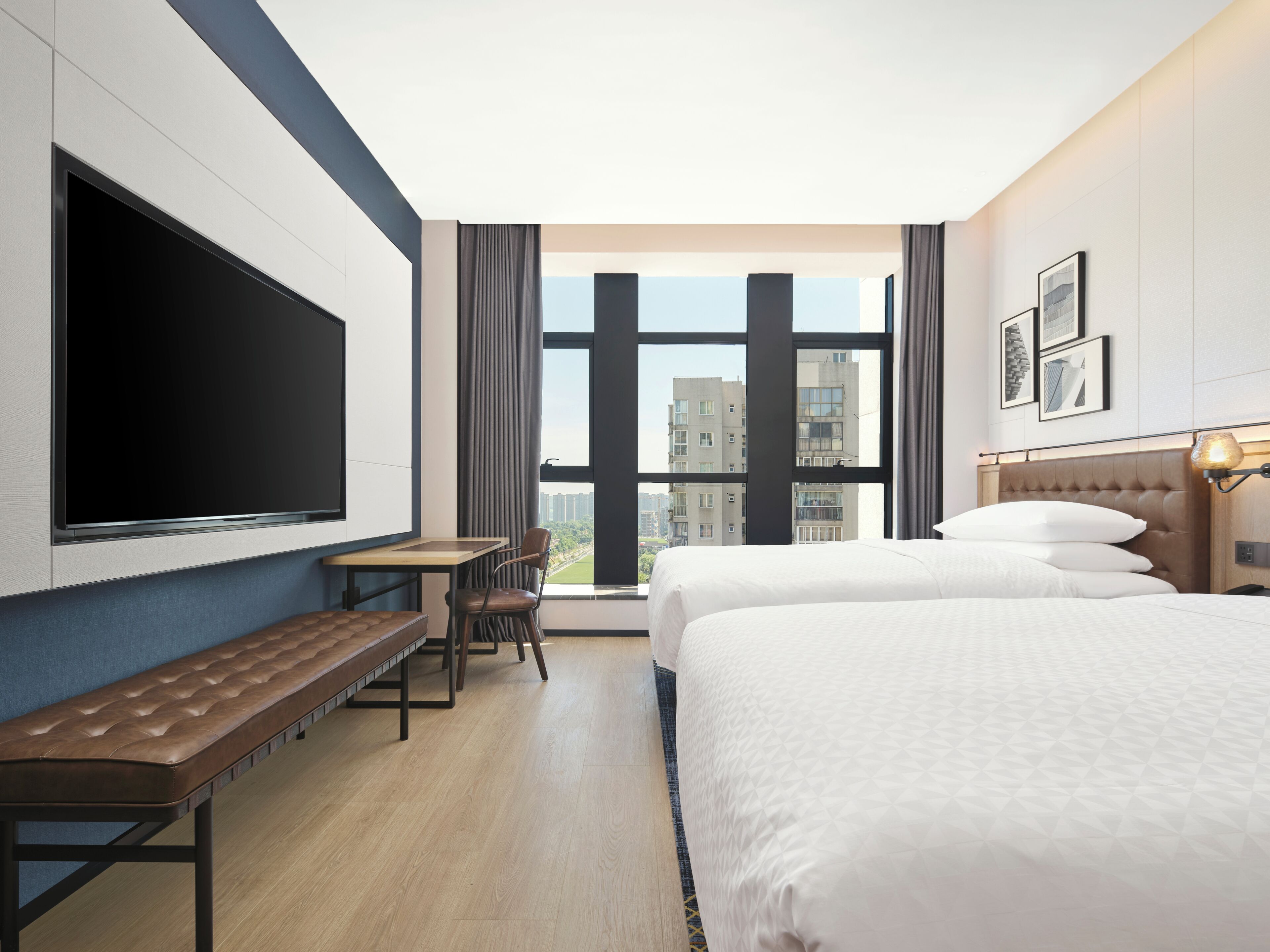 Une chambre d’hôtel équipée d’une grande télévision, d’un bureau, de deux lits et offrant une vue sur les bâtiments par la fenêtre.