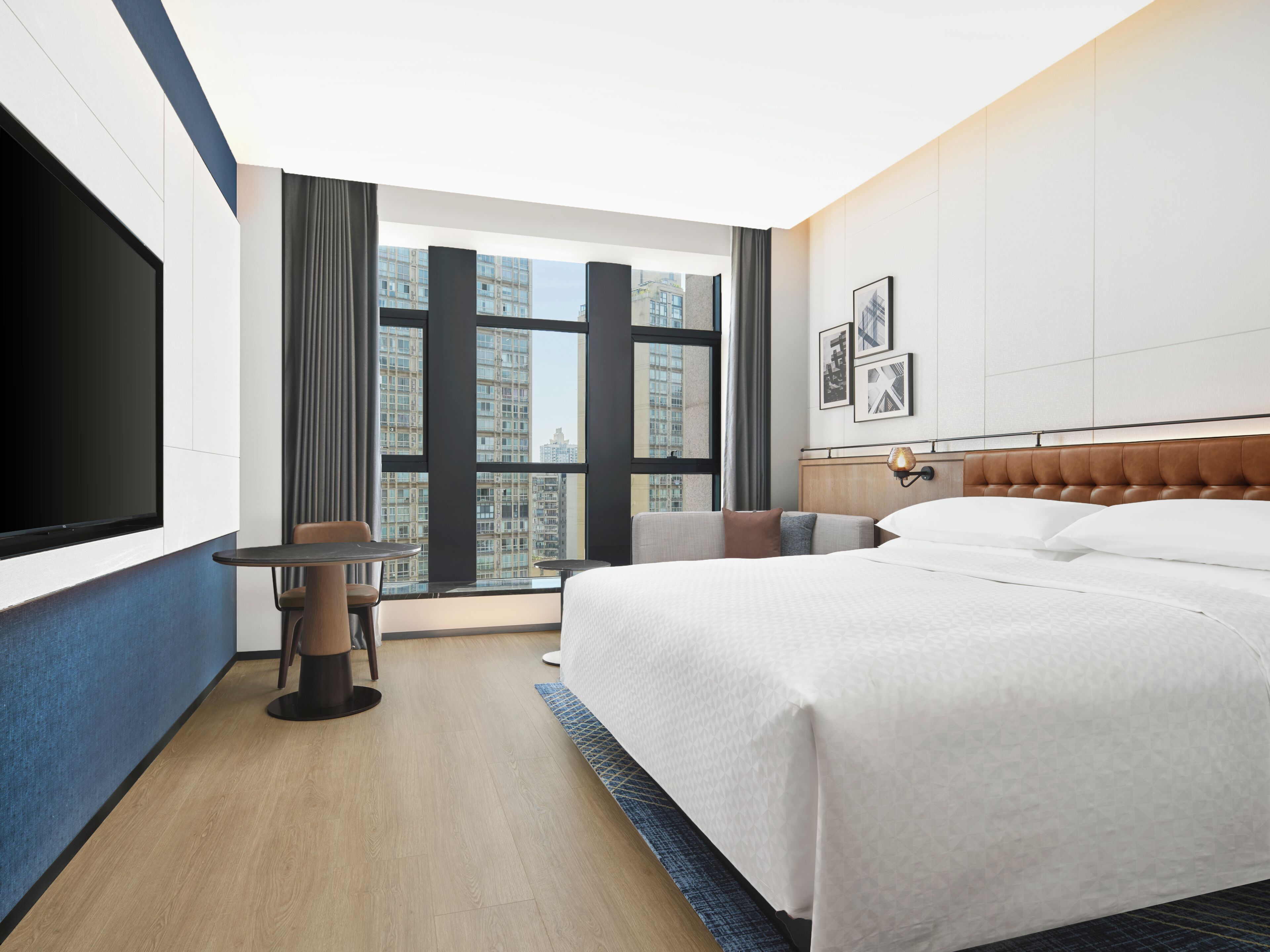 A hotel room with a large bed, a TV, a small table, and a view of high-rise buildings.