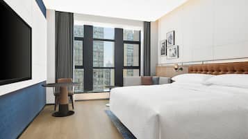 A hotel room with a large bed, a TV, a small table, and a view of high-rise buildings.