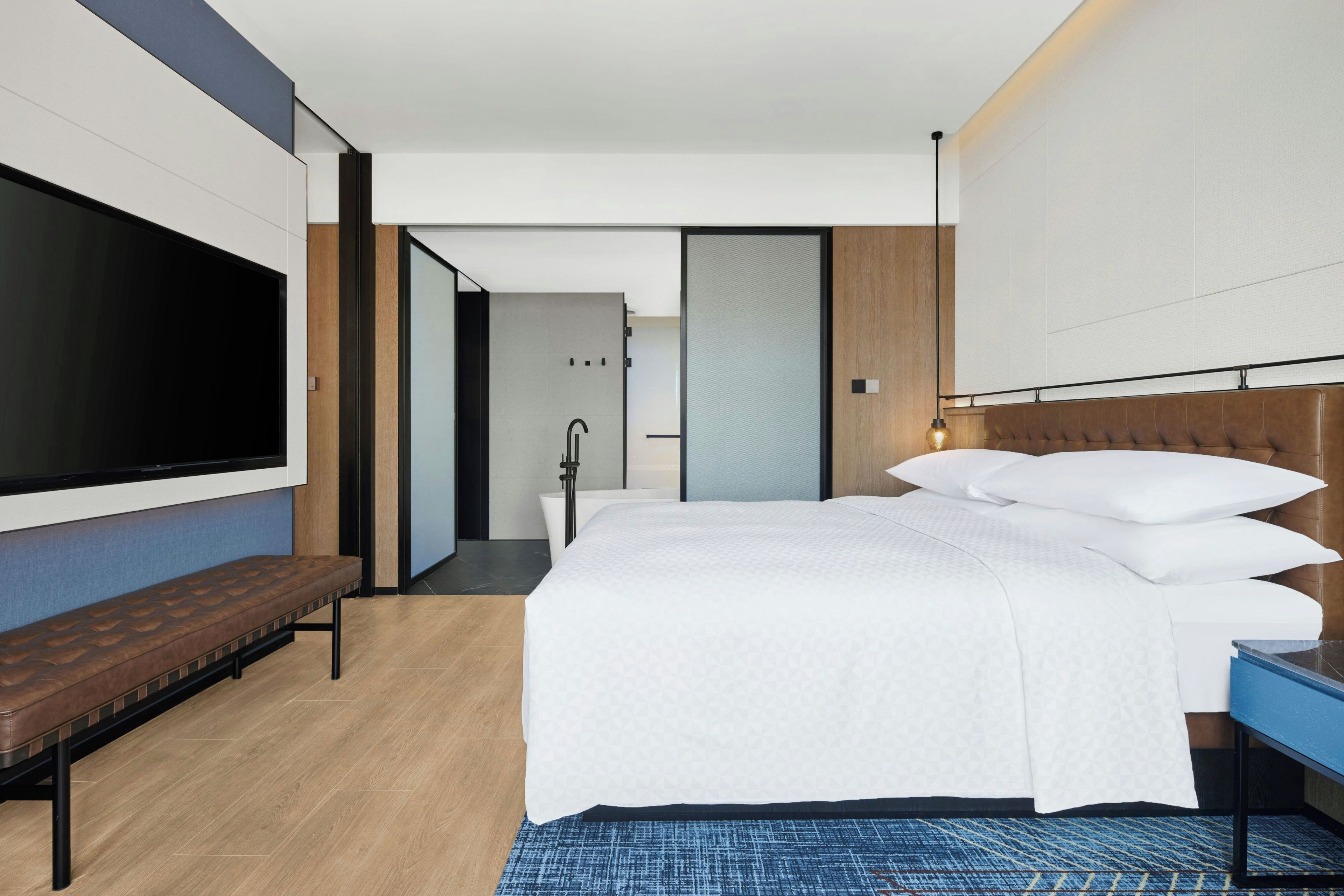 A modern hotel room with a large bed, a bench, a TV, and a bathroom visible through an open door.