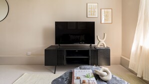 A modern living room with a flat-screen TV on a black entertainment unit, a book titled 'CREATE AT HOME WITH OLD & NEW' on a marble-patterned ottoman, and two framed artworks on the wall. - Take Five on the Green (London)