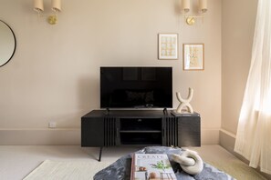 A modern living room with a flat-screen TV on a black entertainment unit, a book titled 'CREATE AT HOME WITH OLD & NEW' on a marble-patterned ottoman, and two framed artworks on the wall. - Take Five on the Green (London)