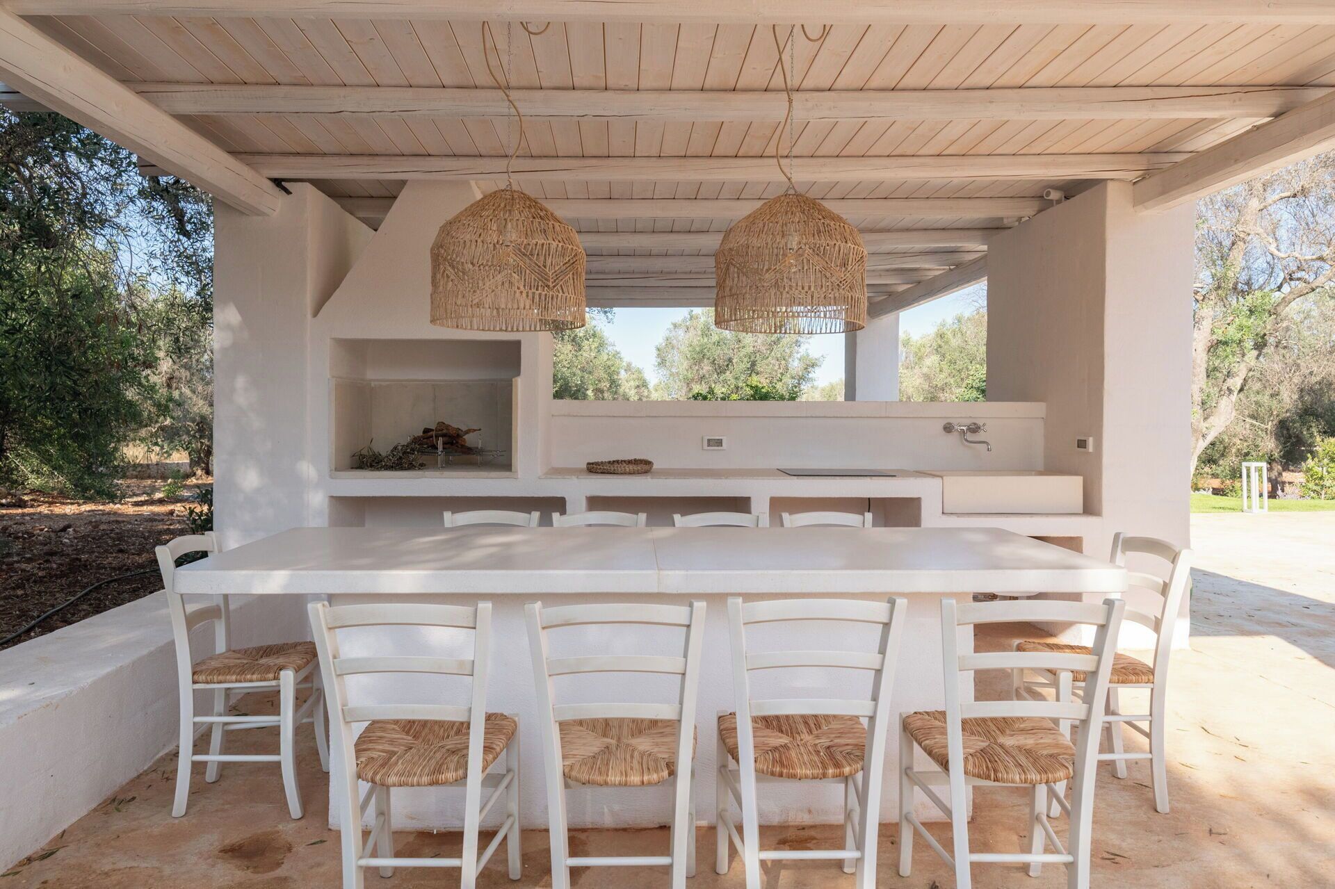 An outdoor dining area with a white table and chairs, two hanging wicker lamps, and a built-in sink.