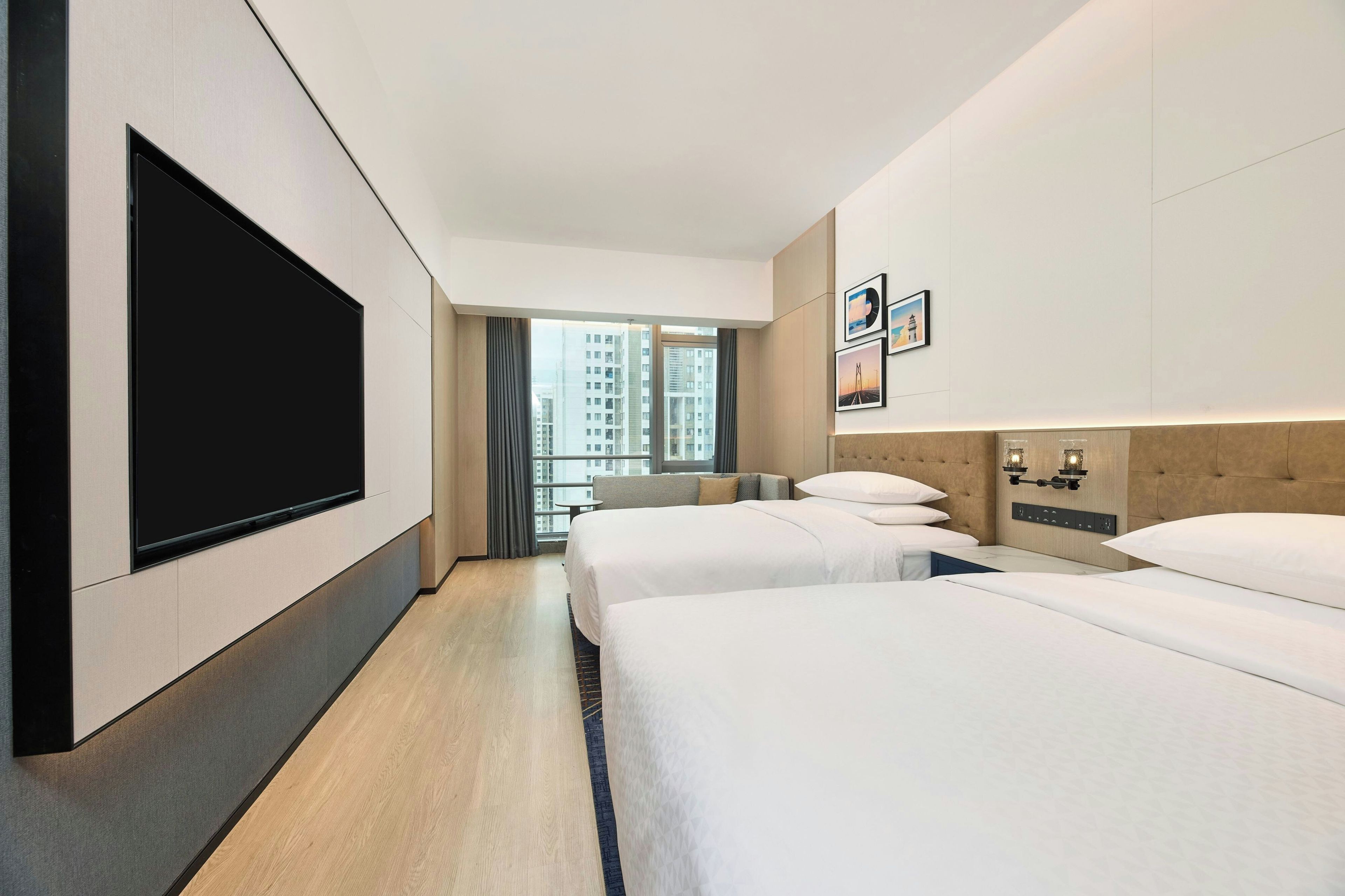 A hotel room with a large flat-screen TV, two beds, a small table, and a view of the cityscape.