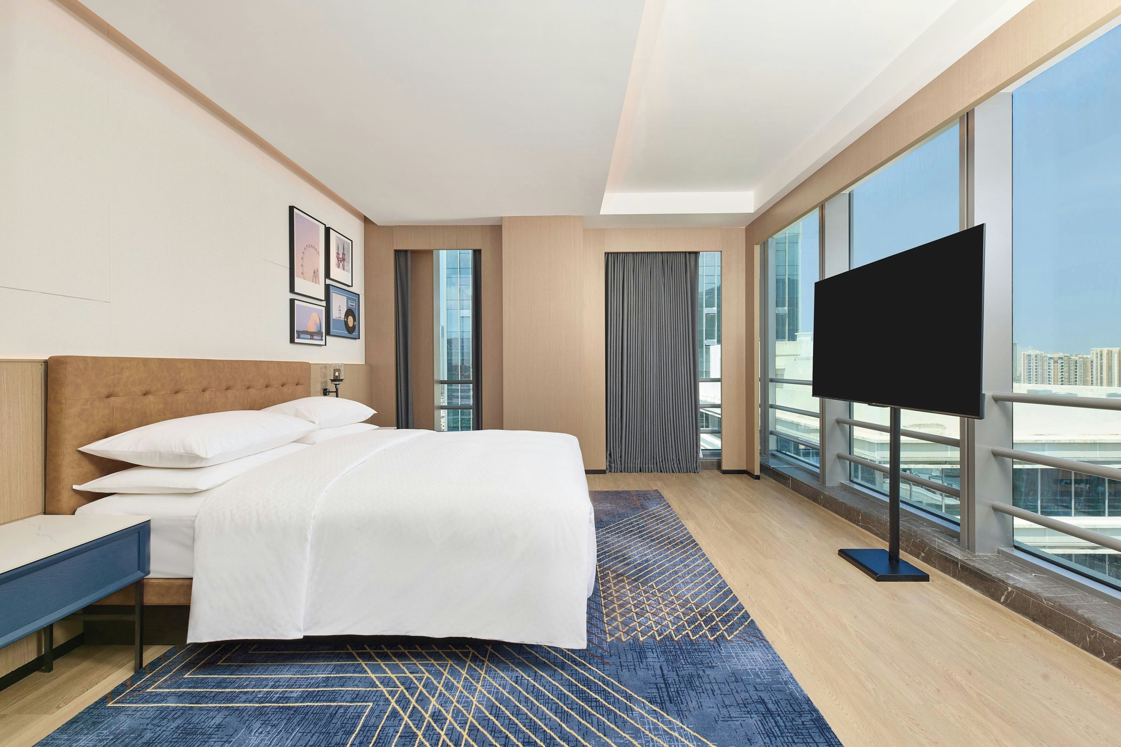 Habitación de hotel con una cama grande, televisor de pantalla plana, escritorio y vistas a la ciudad a través de amplios ventanales.
