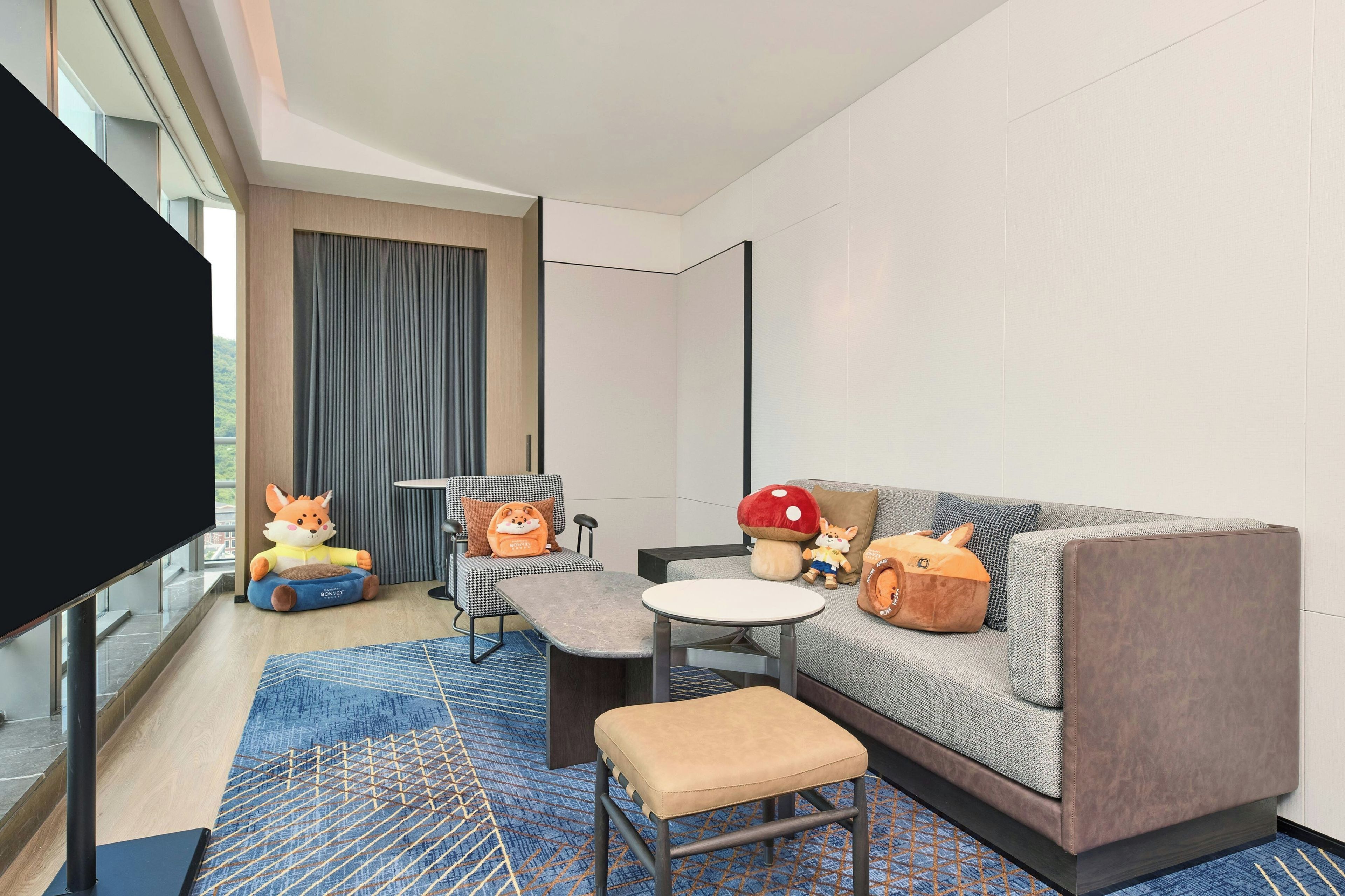 A modern hotel room with a sofa, a small table, a flat-screen TV, and a window with curtains.