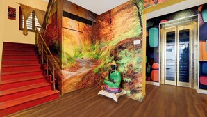 A staircase with red steps leading to a landing, a wall with a mural, and a statue of a seated figure. - Collection O Omni Bus Terminal (Coimbatore)