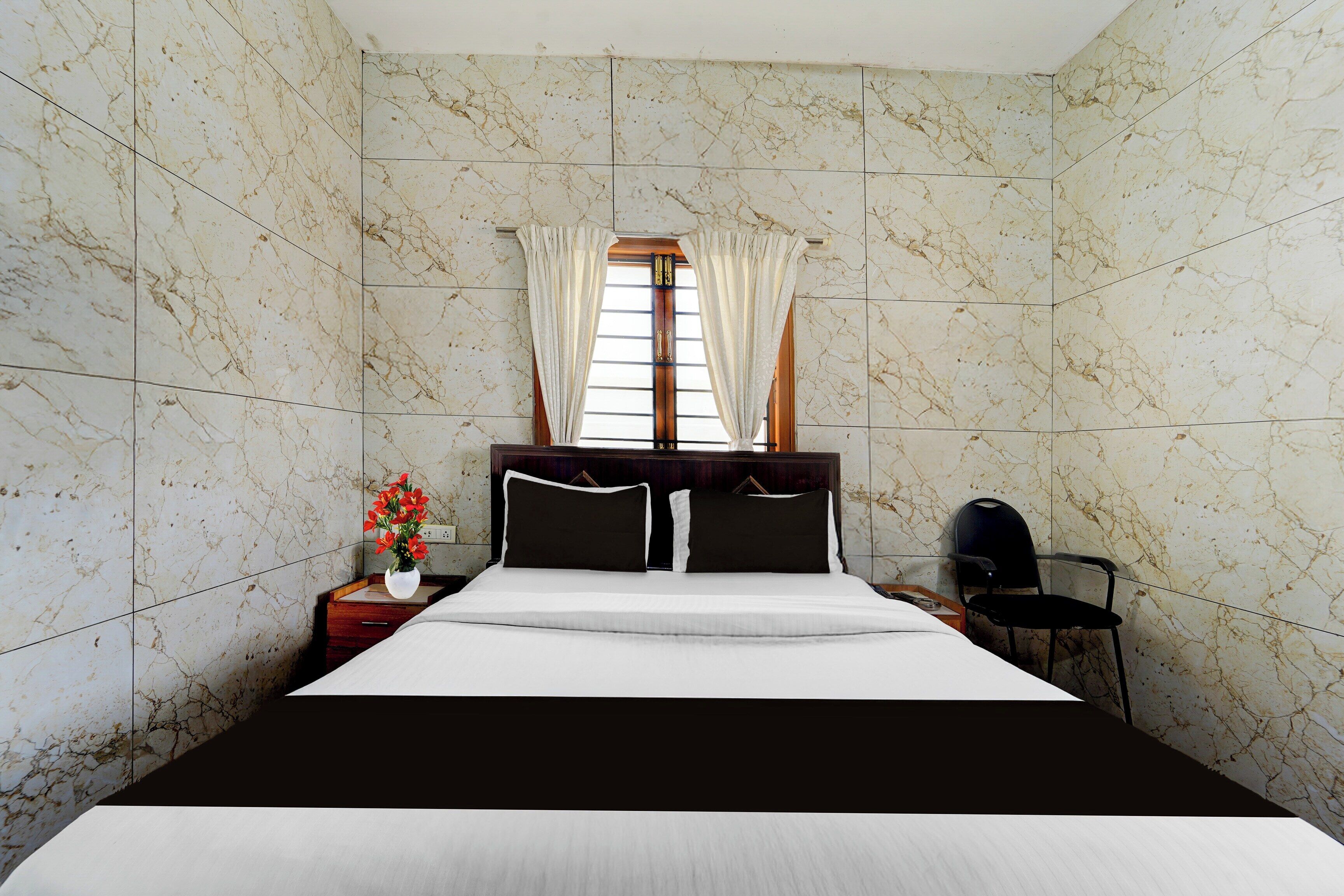 A hotel room with a marble-patterned wall, a bed with black and white bedding, a nightstand with a vase of flowers, and a chair.