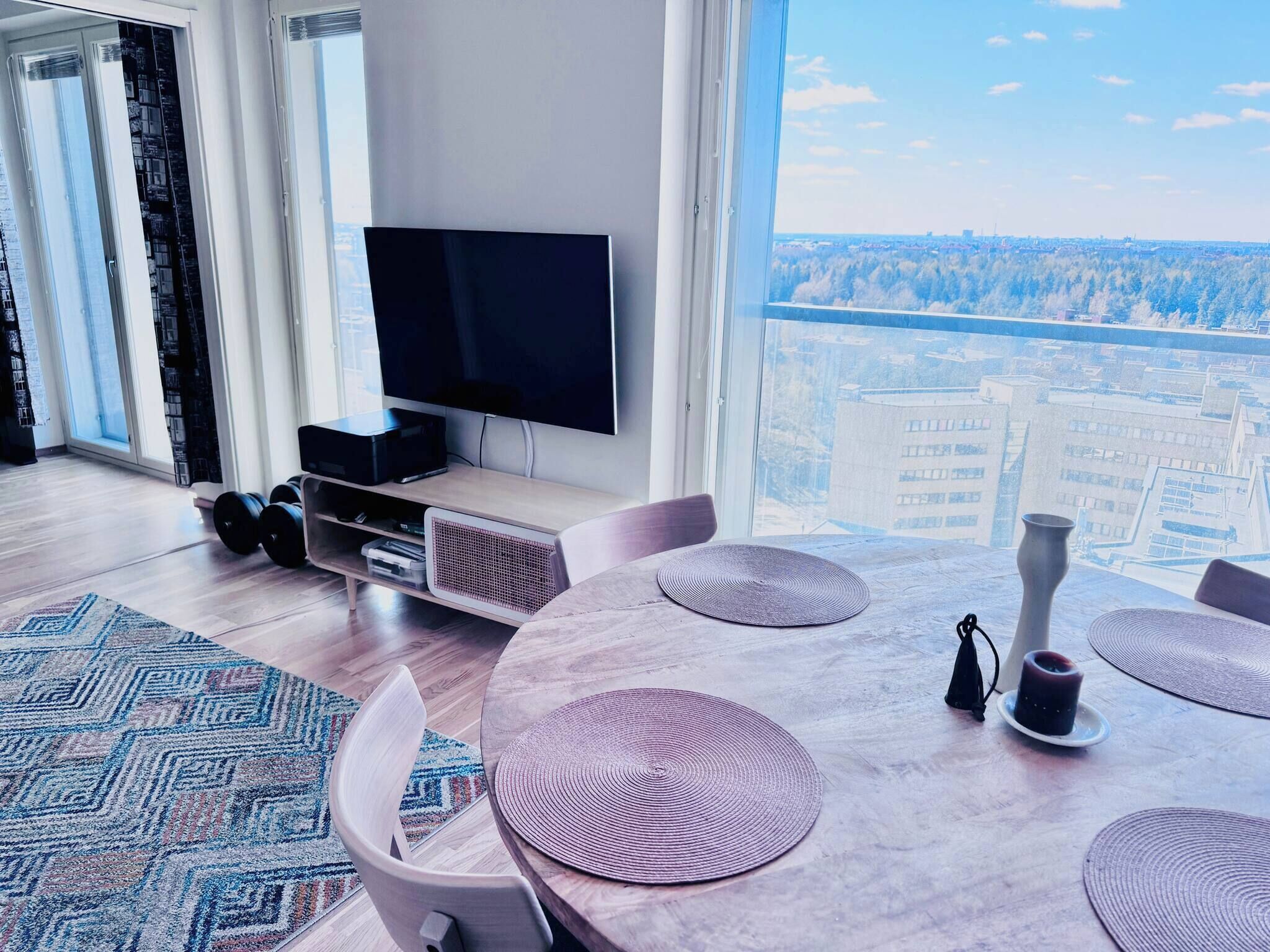 A modern living room with a flat-screen TV, a dining table with placemats, and a view of a cityscape through large windows.