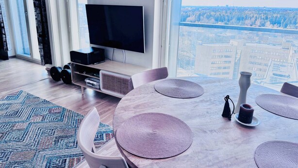 Uma sala de estar moderna com uma televisão de tela plana, uma mesa de jantar com jogos americanos e vista para o horizonte urbano através de grandes janelas.