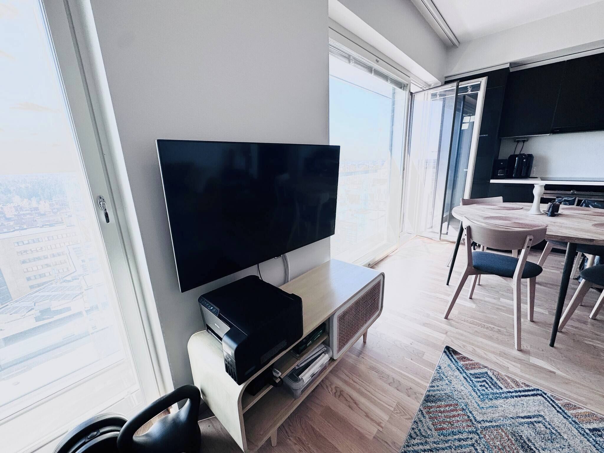 A modern living room with a flat-screen TV, wooden flooring, and a large window offering a city view.