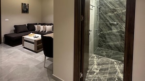 A modern living room with a sofa, coffee table, and a glass door leading to a shower area with marble tiles. - 304 Al-najeh JRE Apartments For Rent ave (Amman)