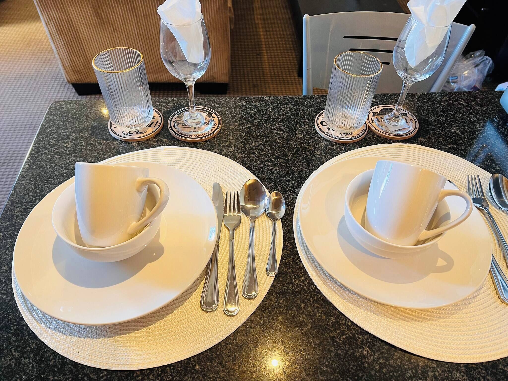 A table set with white plates, cups, cutlery, and napkins on a granite countertop.