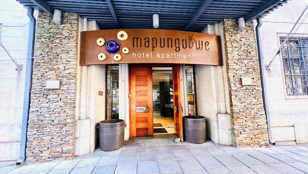 Entrada de un hotel con fachada de piedra, un letrero que dice 'Mapungubwe Hotel Apartments' y dos contenedores de basura.
