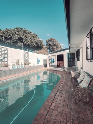 Una piscina con solárium de ladrillo, mobiliario exterior y un edificio blanco con ventanas de celosía.