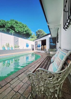 A pool area with a bench, a covered patio, and a building with a garage door. - Bonita Vista Plattekloof (Cape Town)