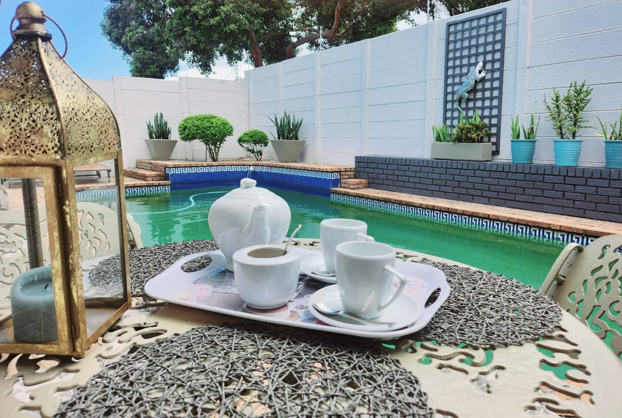 A poolside setting with a tray of white cups and a teapot, surrounded by greenery and a white wall.