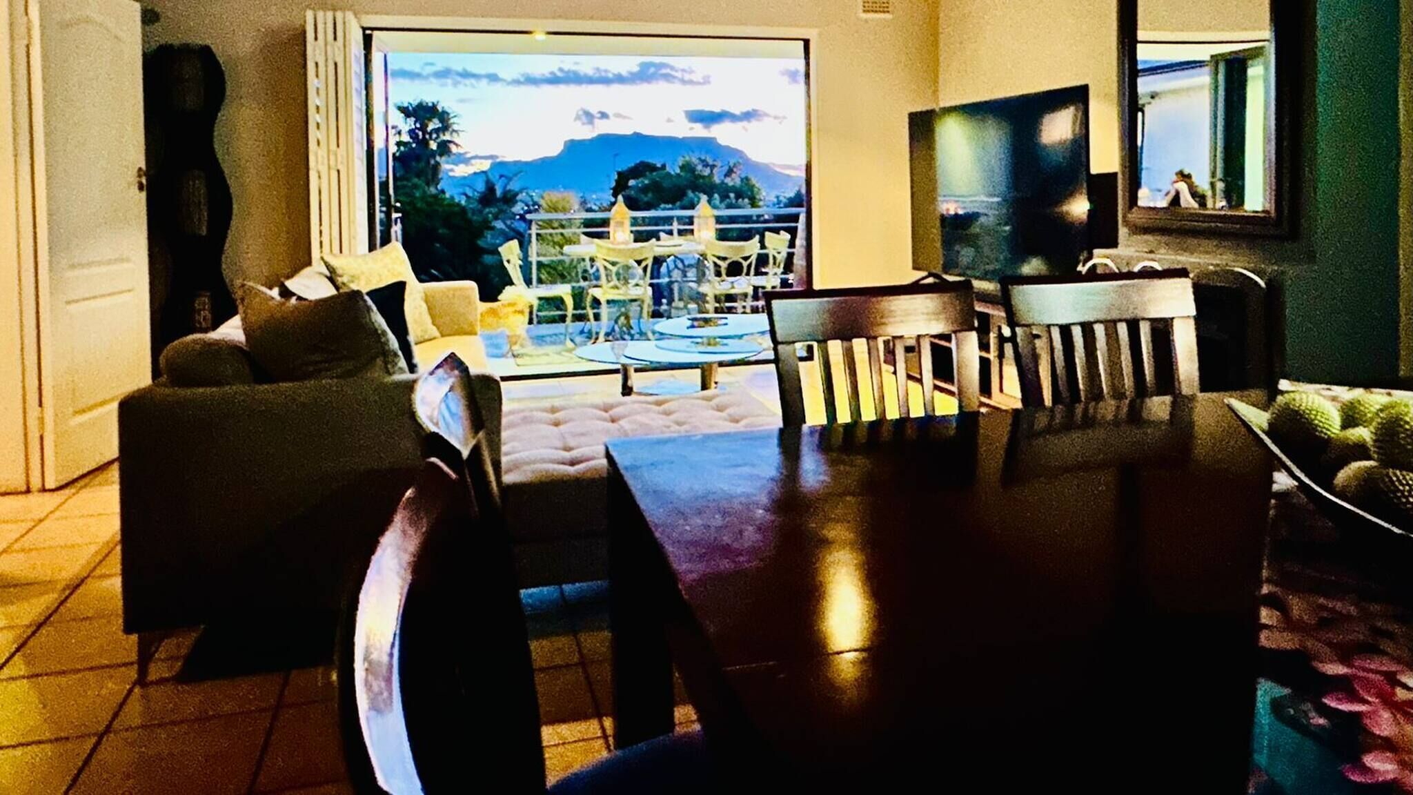 A living room with a sofa, a dining table, and chairs. There is a TV on the wall and a view of the outdoors.