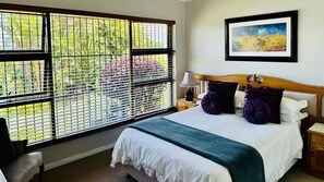 A hotel room with a large window, a bed with white linens, purple pillows, a wooden headboard, a nightstand with a lamp, and a framed painting on the wall. - Bonita Vista Plattekloof (Cape Town)