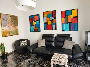 Ein modernes Wohnzimmer mit einer schwarzen Ledersofa, zwei gemusterten Zierkissen, einem Marmorboden und drei abstrakten Bildern an der Wand.