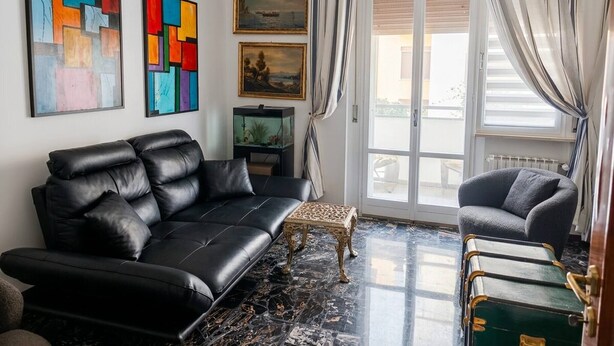 A modern living room with a black leather sofa, a small table, and a green trunk. There are colorful abstract paintings on the wall and a window with curtains. - Modern Apartment Near Senigallia's Velvet Beach (Senigallia)