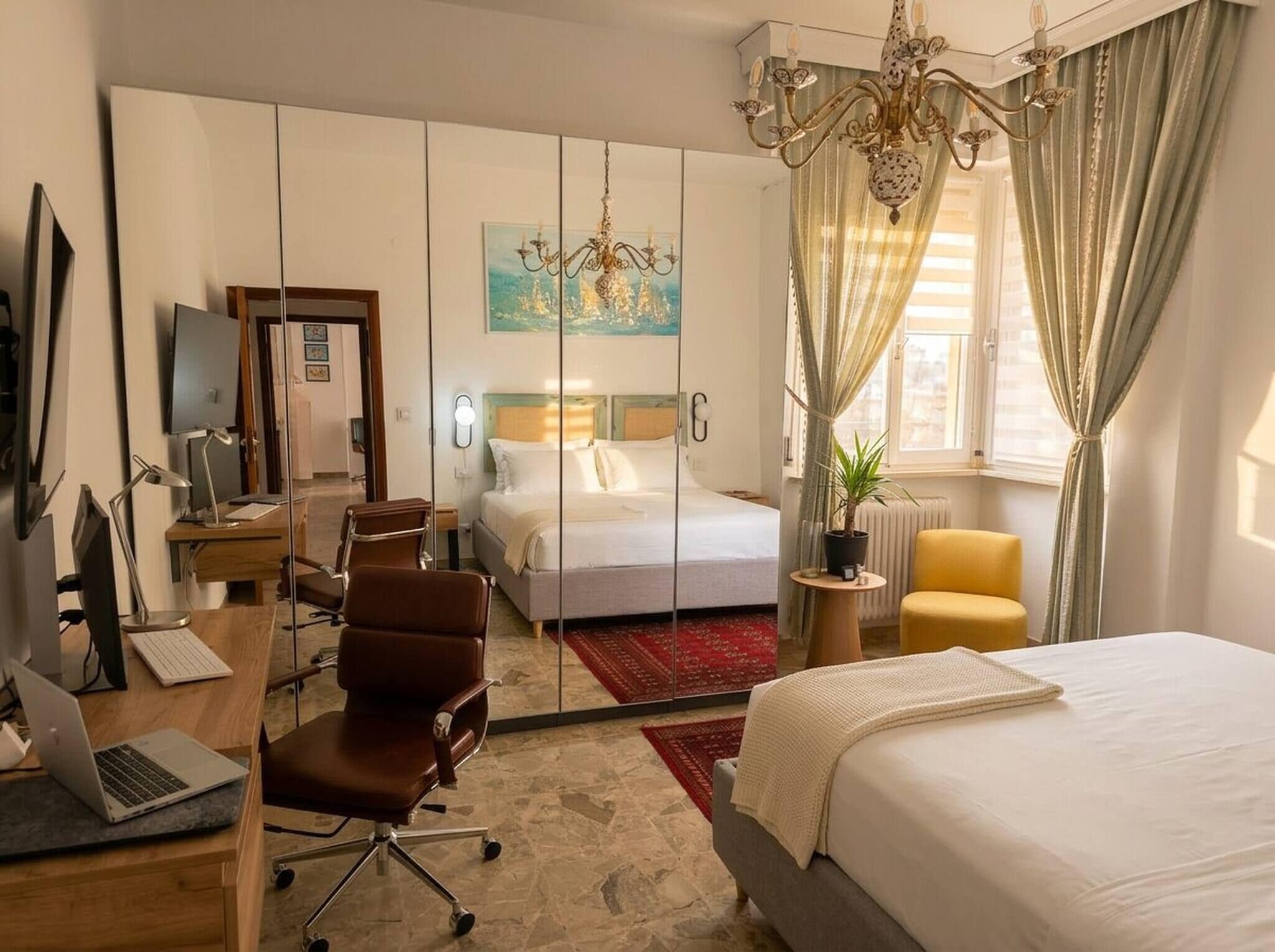 A hotel room with a bed, desk, chair, computer, TV, and a painting on the wall.