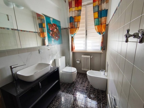 A bathroom with a white sink, a toilet, and a bidet. There is a mirror above the sink and a window with colorful curtains. - Modern Apartment Near Senigallia's Velvet Beach (Senigallia)