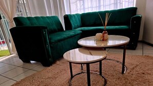 Una sala de estar con un sofá verde, dos mesas redondas, un felpudo mullido y una ventana con cortinas.