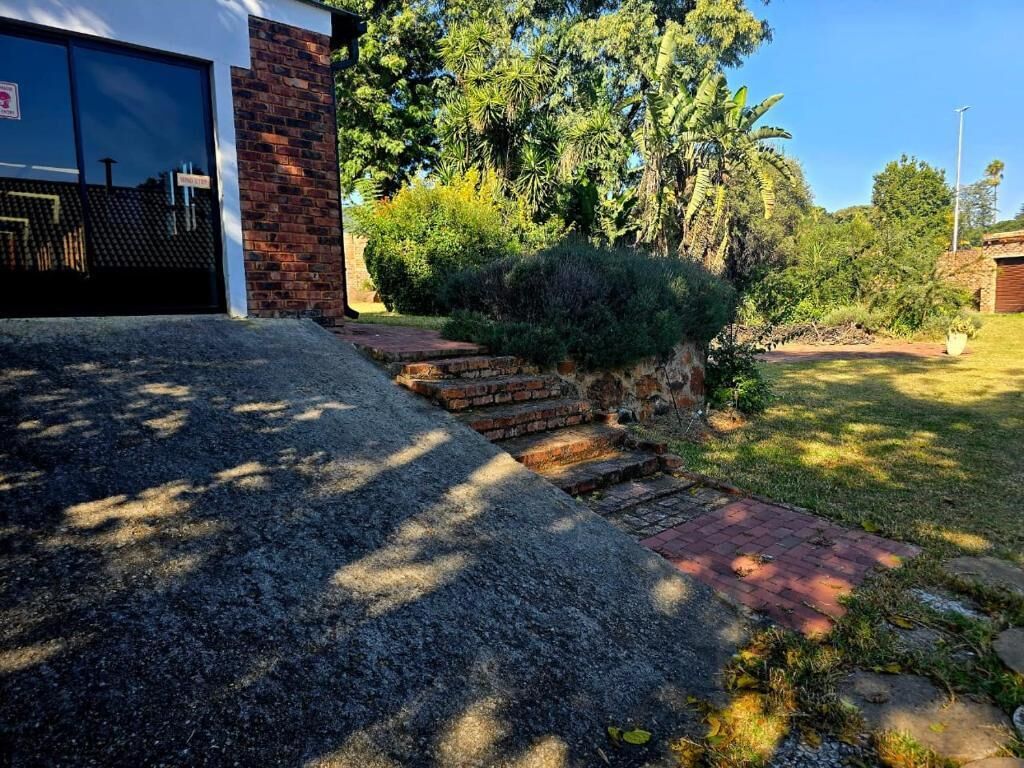 Um imóvel residencial com um acesso pavimentado, um caminho de tijolos que leva a uma garagem e um jardim exuberante com diversas plantas e árvores.