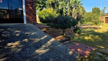 Um imóvel residencial com um acesso pavimentado, um caminho de tijolos que leva a uma garagem e um jardim exuberante com diversas plantas e árvores.