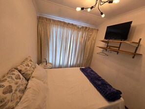 A bedroom with a bed, curtains, a TV on a wall-mounted shelf, and a bedside table. - Room-aquasaintsguest Lodge (Johannesburg)