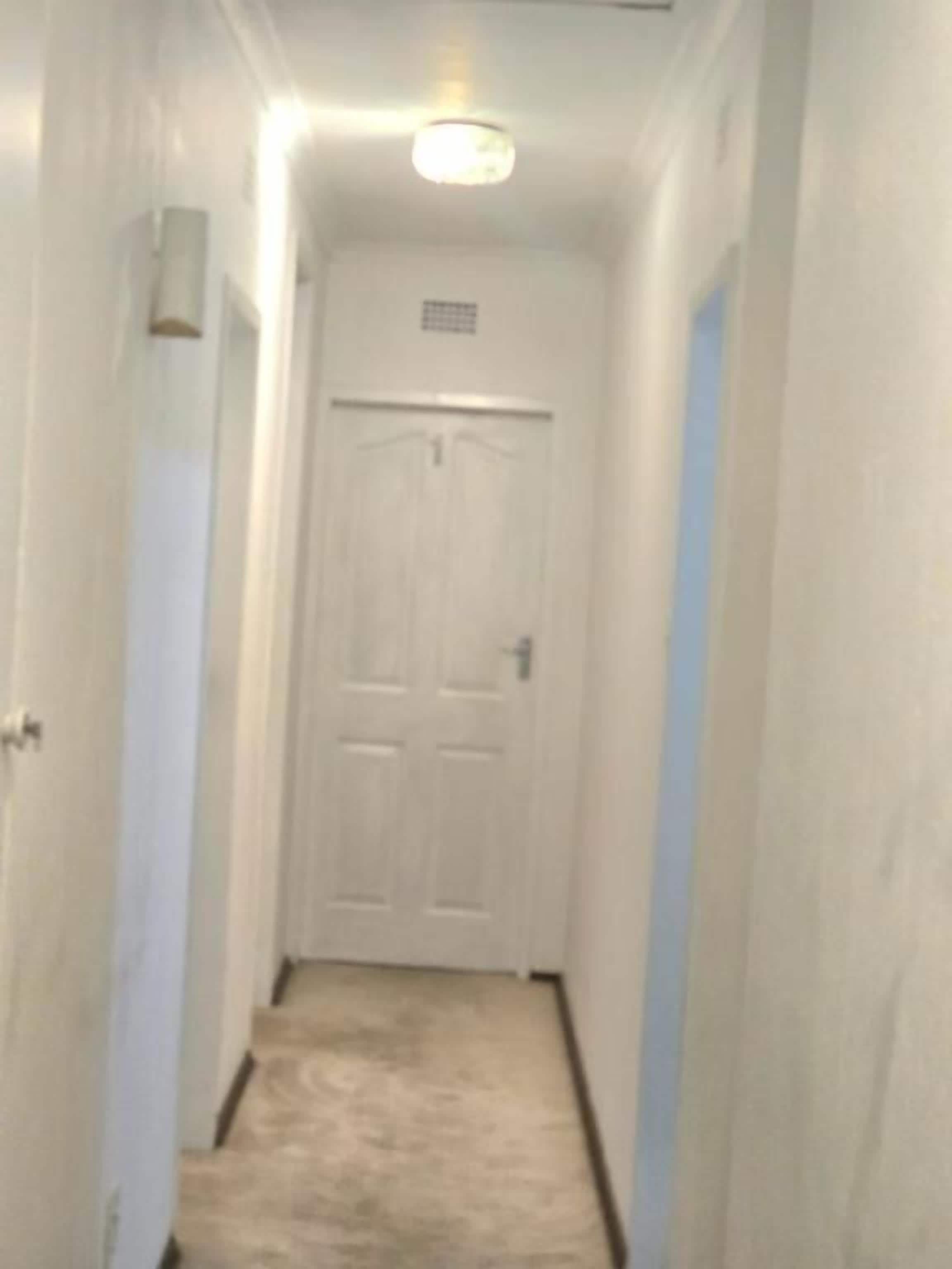 A narrow hallway with a white door at the end, a light fixture above, and a carpeted floor.