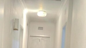 Un pasillo estrecho con una puerta blanca al final, una lámpara arriba y piso cubierto de alfombra.