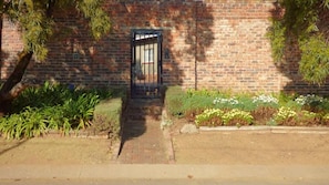 Un edificio de ladrillo con una entrada porticada, un sendero pavimentado que lleva a la puerta, y un jardín con diversas plantas y flores.
