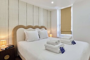 A hotel room with a bed, pillows, a bedside lamp, a window with a blind, and a stack of towels. - Apartment in the heart of the Marais (Paris)