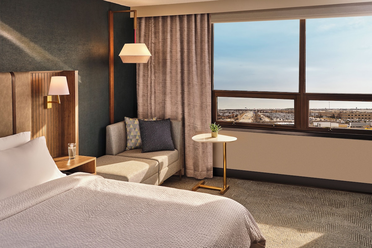 Habitación de hotel con una cama grande, una mesita de noche, una lámpara, una pequeña mesa con una planta, un sofá con cojines, una ventana con vistas a la ciudad y una cortina.