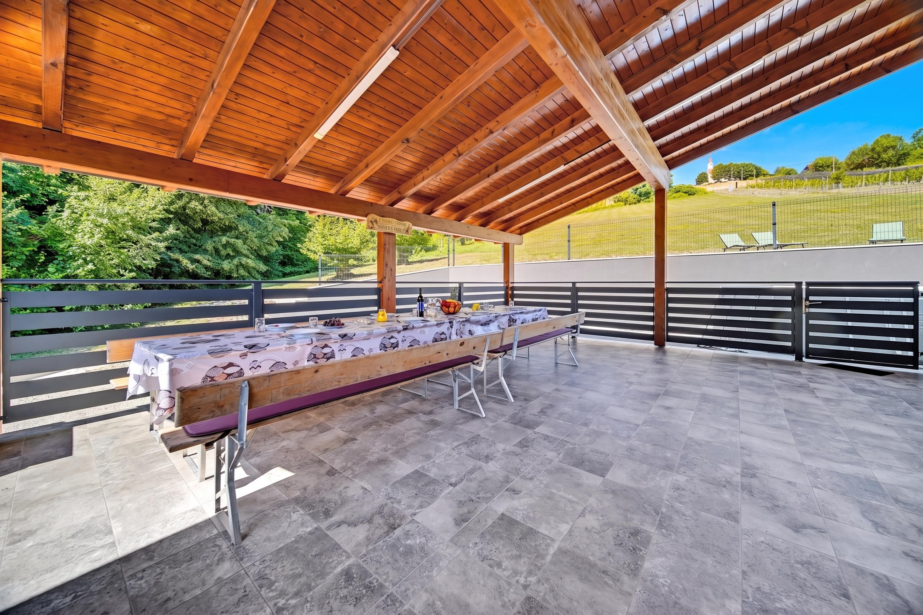 An outdoor covered patio with a long dining table set for a meal, surrounded by metal railings and a view of a green landscape.
