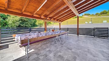 Un patio techado al aire libre con una larga mesa de comedor dispuesta para una comida, rodeado de barandillas metálicas y vistas a un paisaje verde.