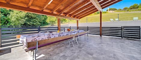 Un patio techado al aire libre con una larga mesa de comedor dispuesta para una comida, rodeado de barandillas metálicas y vistas a un paisaje verde.
