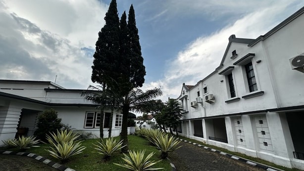 A courtyard with a tall coniferous tree, a white building with multiple windows, and a clear sky. - Casa D'Ladera Bandung Standard Double (Bandung)