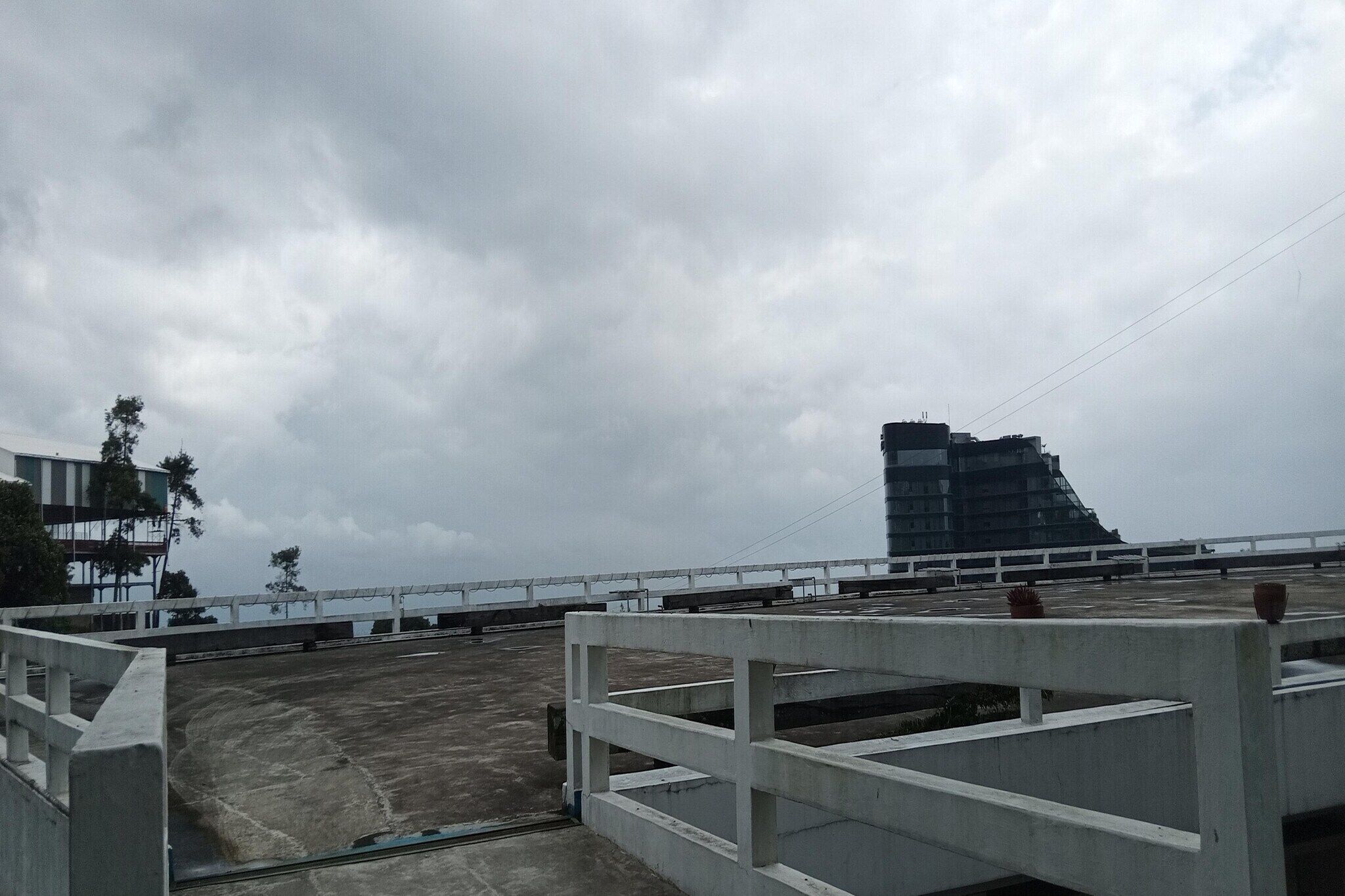 A rooftop with a concrete barrier, a building in the background, and a cloudy sky.