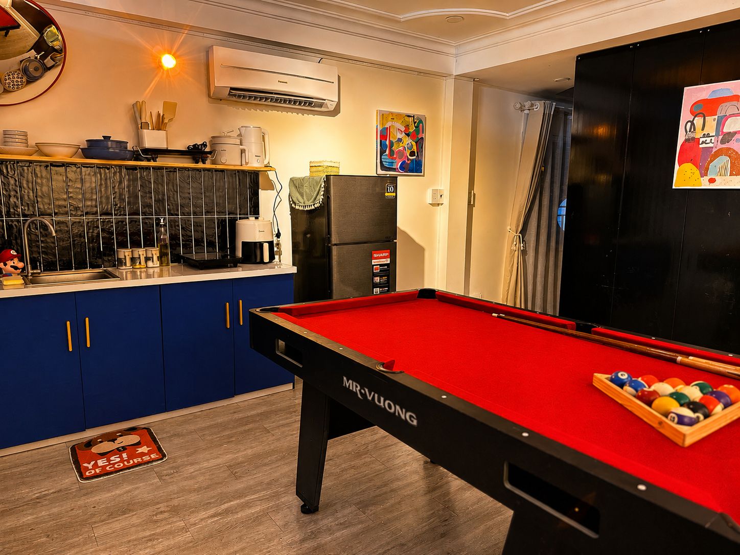 A pool table with a red felt surface and a cue stick, a refrigerator, a microwave, and a sink with various kitchen items.