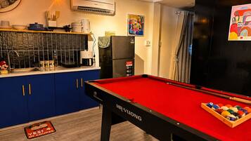 A pool table with a red felt surface and a cue stick, a refrigerator, a microwave, and a sink with various kitchen items.