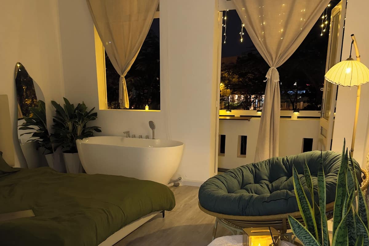 Un dormitorio moderno con un ventanal grande, una bañera, un sofá verde y una planta en maceta.