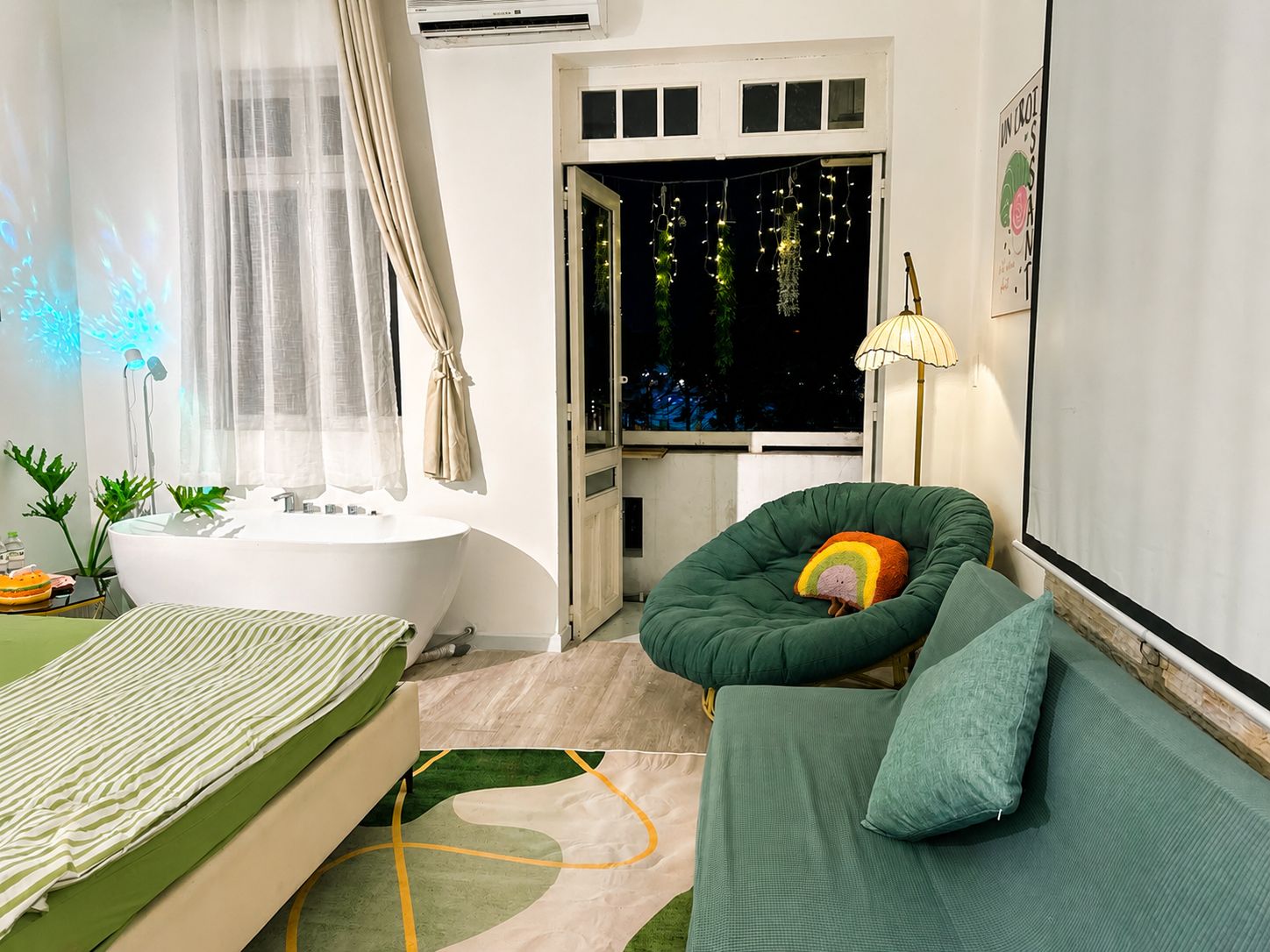 Ein modernes Schlafzimmer mit einem grünen Sofa, einem Bett mit gestreifter Bettwäsche, einer weißen Badewanne und einem Fenster mit Lichterketten.