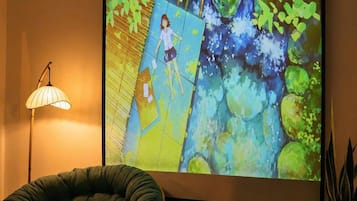 Una habitación con una pantalla grande mostrando una escena animada, un sillón de felpa verde, una mesita con una vela y una planta en maceta.