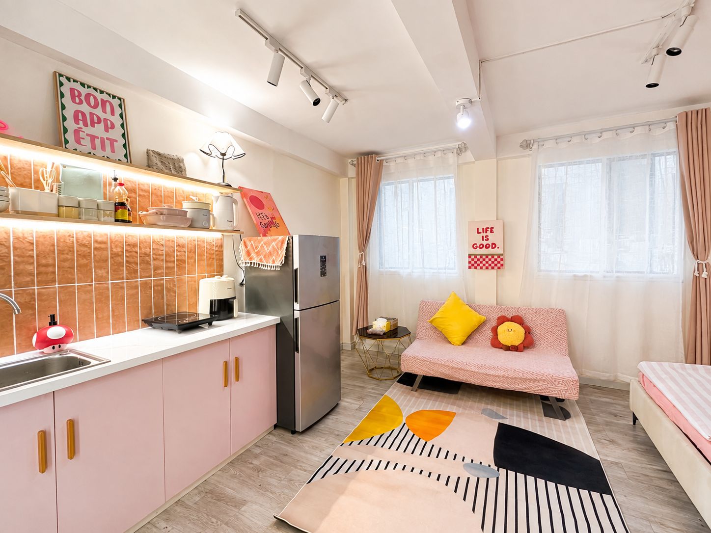 Una cocina moderna con refrigerador, cafetera y lavamanos. Hay un sofá con cojines y una almohada decorativa. La habitación tiene un ventanal con cortinas y una pared con una cita inspiradora.