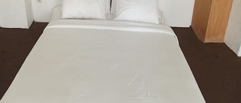 Una cama con ropa de cama blanca en una habitación con alfombra marrón y paredes blancas.