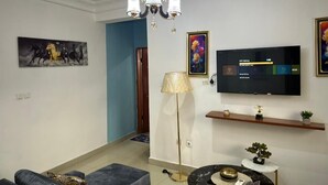 A living room with a sofa, a coffee table, a TV, a chandelier, and a painting on the wall. - Comfy Cityscape Elegance (Yaoundé)