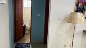 A hallway with a blue wall, a wooden door leading to a bathroom, a standing lamp, and a blue mat on the floor. - Comfy Cityscape Elegance (Yaoundé)